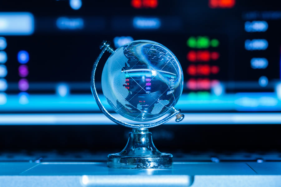 Crystal globe on a silver stand in front of a blurred trading screen showing red and green market prices