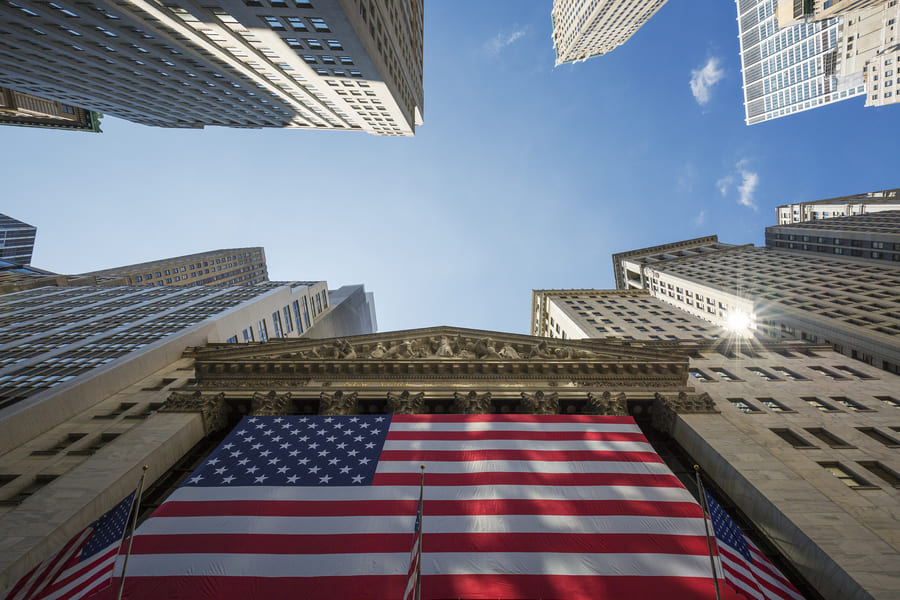 Low-angle view of the New York Stock Exchange facade draped in a large US flag, framed by Wall Street skyscrapers